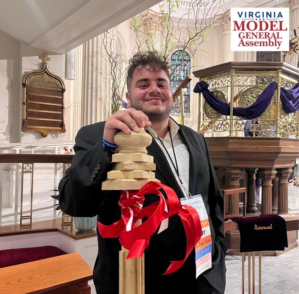 A smiling young man wearing a suit and a multi-ribboned nametag, resting his hand on a wooden ceremonial mace tied with red ribbon, featuring the Virginia Model General Assembly logo.