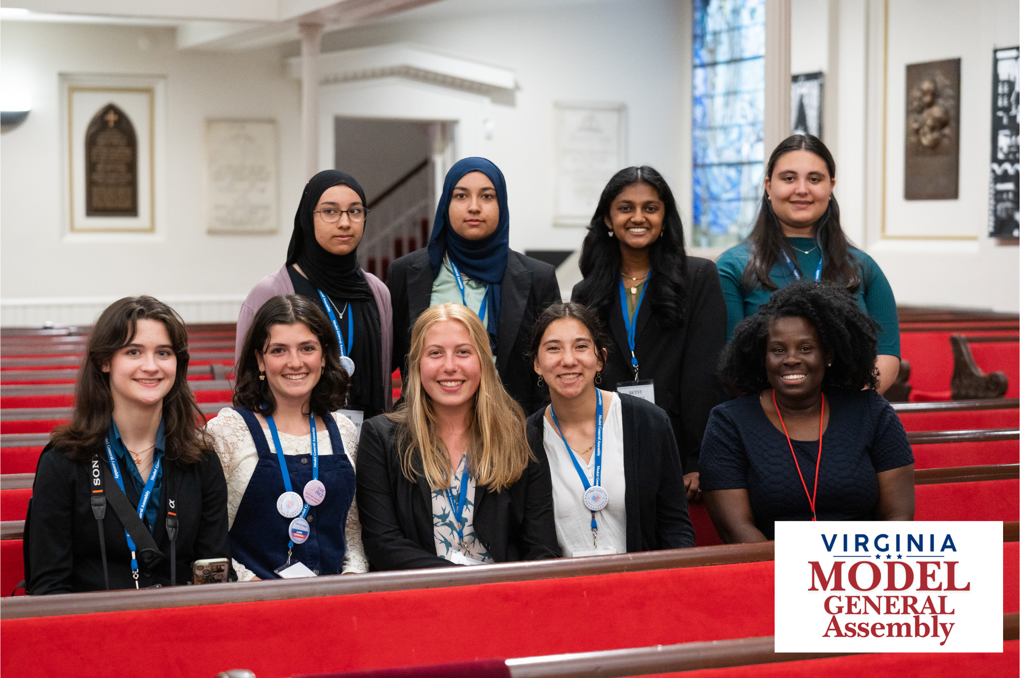 A group photo of nine smiling young women wearing lanyards and posing in red pews, featuring the Virginia Model General Assembly logo.