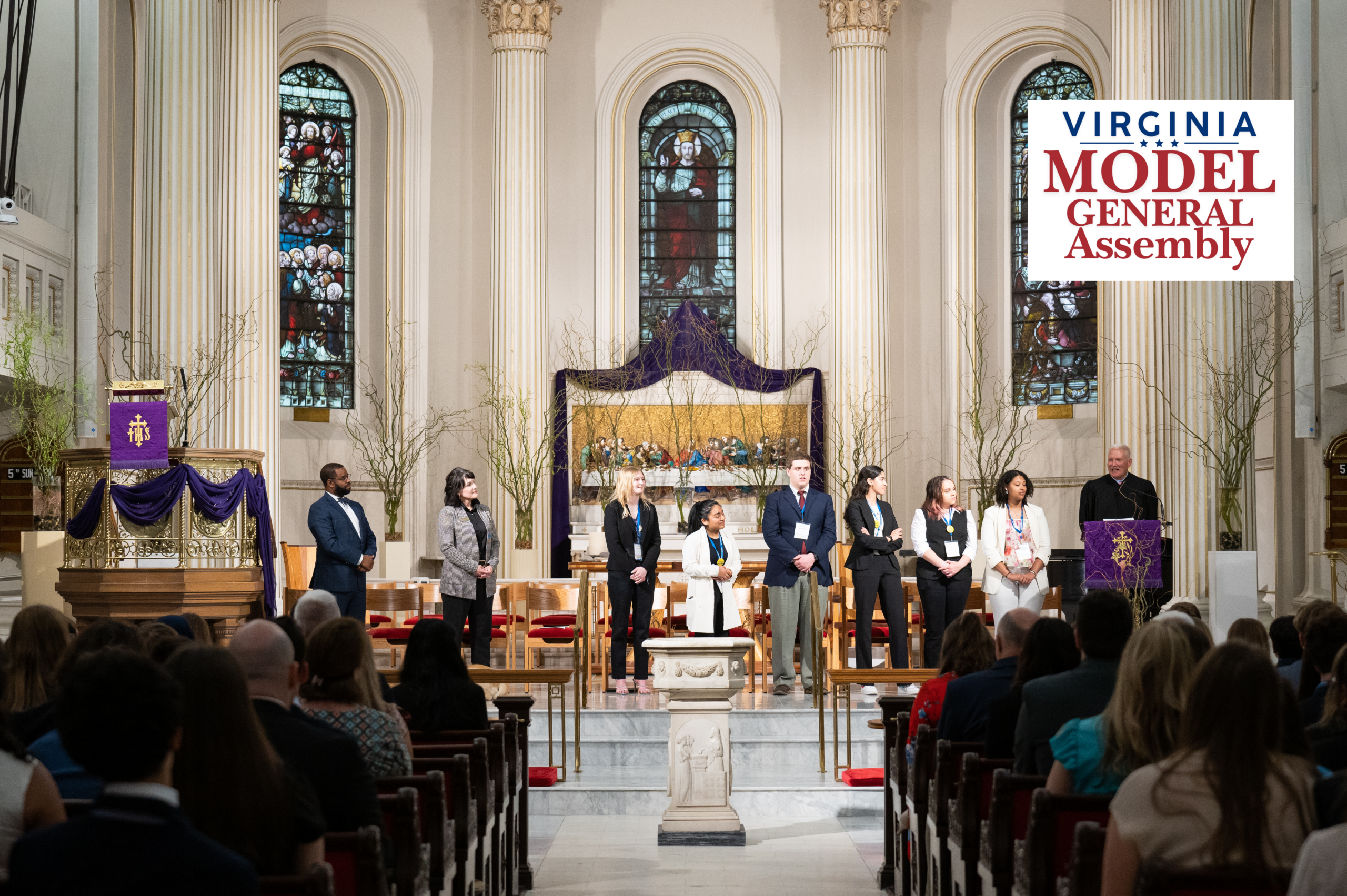 A line of young adults and a speaker standing at the front of a church sanctuary facing an audience seated in pews, featuring the Virginia Model General Assembly logo.