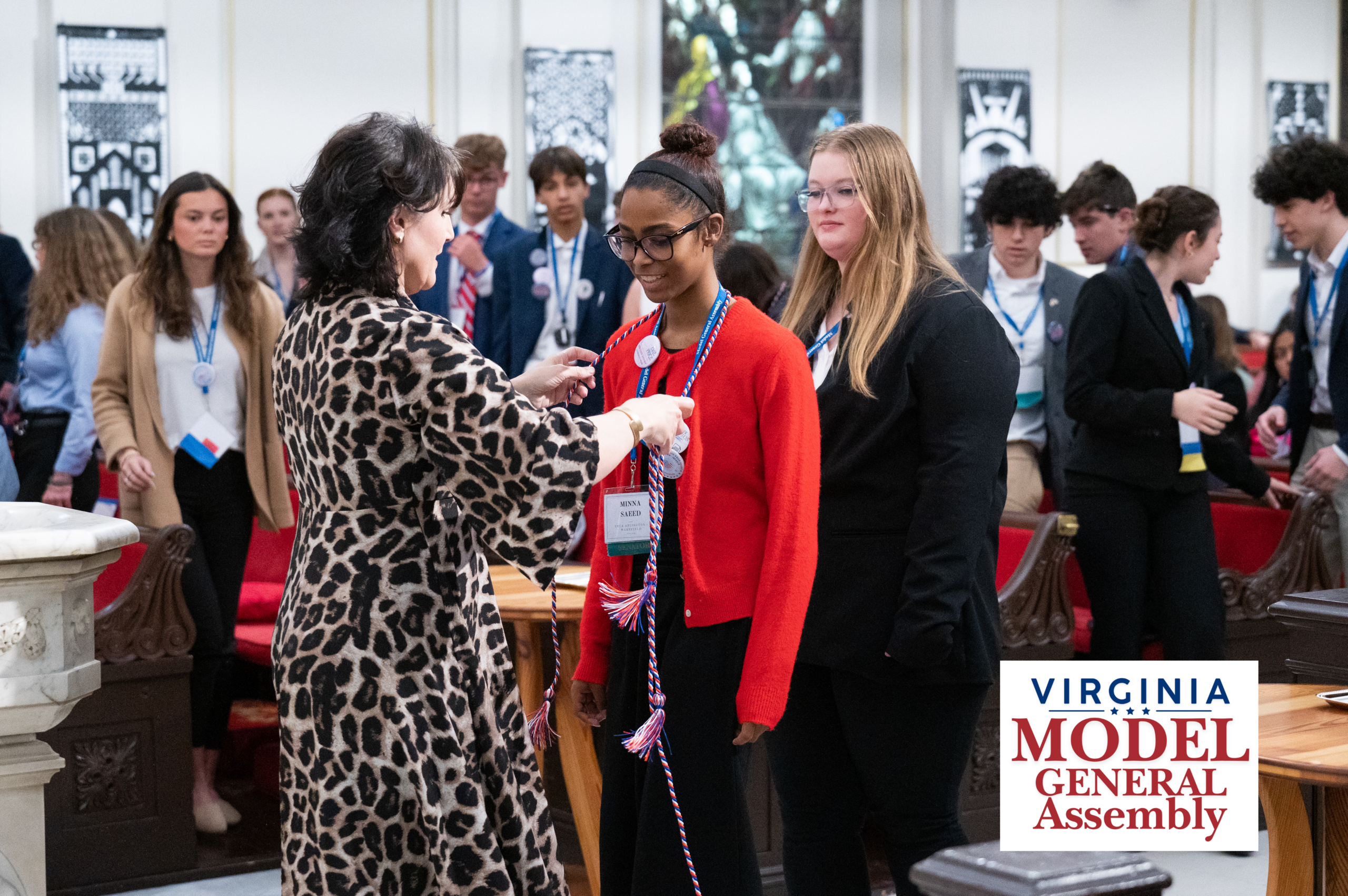 A woman placing a red, white, and blue honor cord around the neck of a smiling young woman, featuring the Virginia Model General Assembly logo.