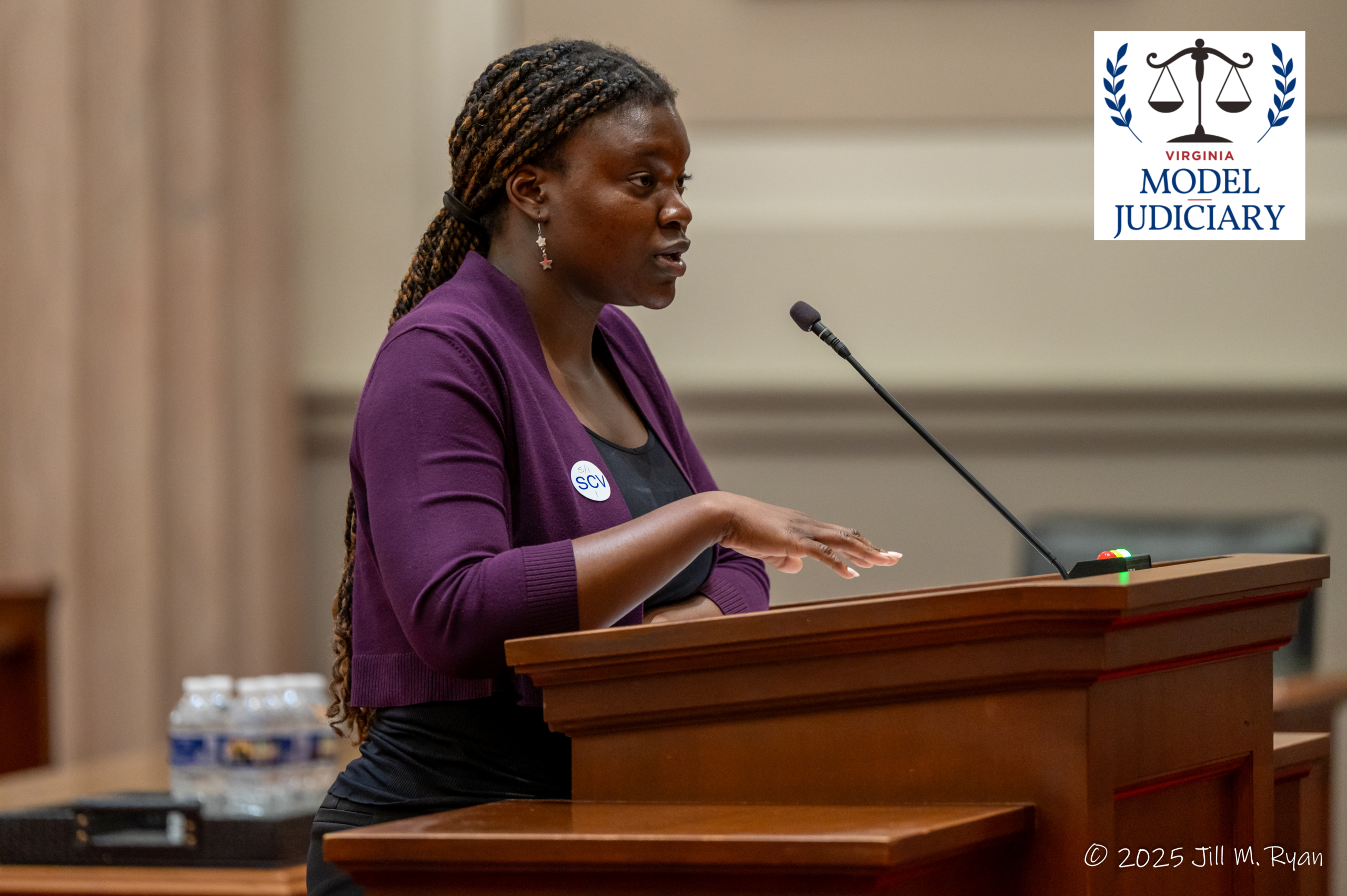 A young woman speaking at a wooden podium with a microphone, featuring the Virginia Model Judiciary logo and a 2025 Jill M. Ryan copyright.