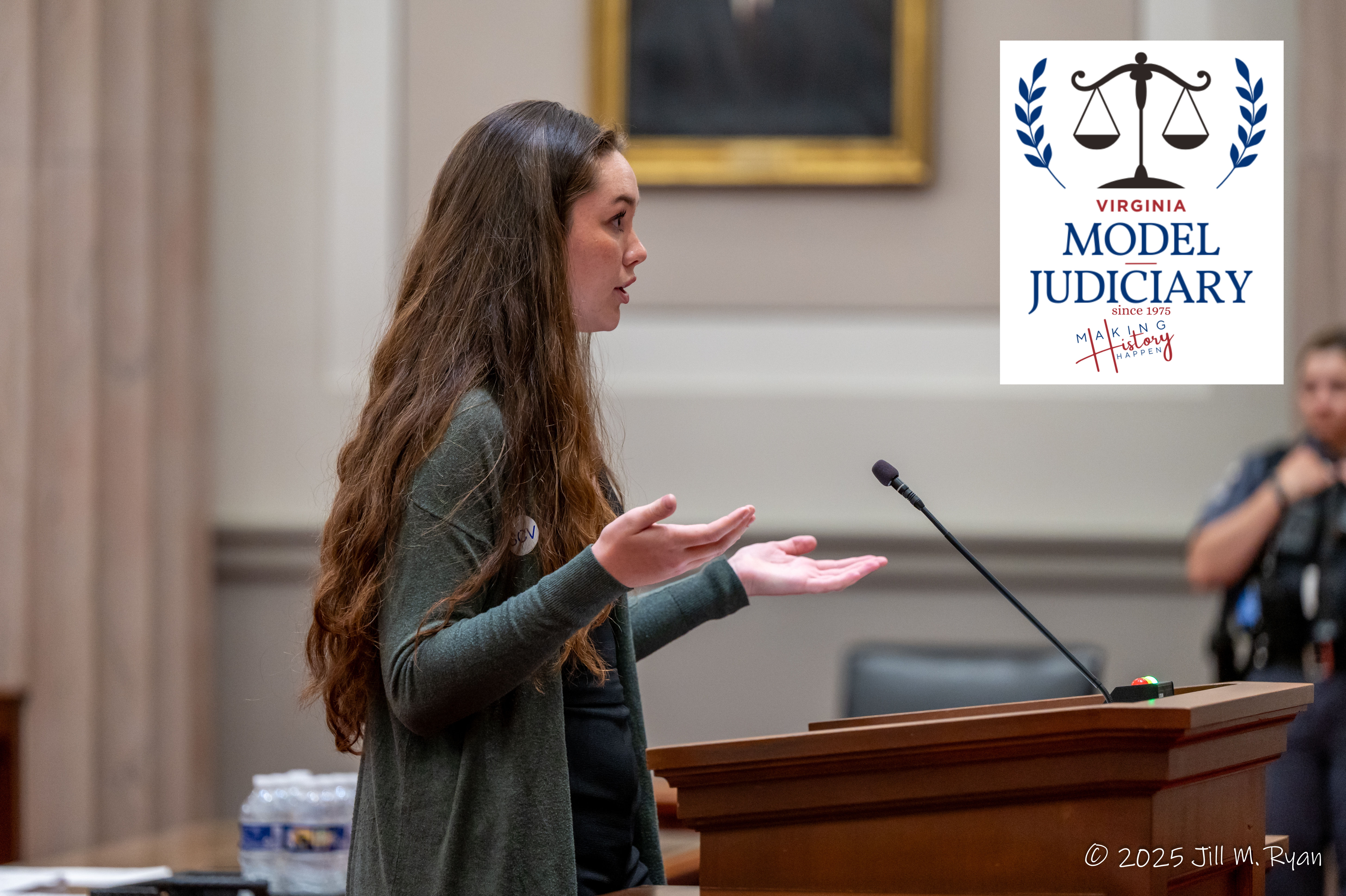 A young woman gesturing with her hands while speaking at a wooden podium, featuring the Virginia Model Judiciary logo with the tagline 'Making History Happen' and a 2025 Jill M. Ryan copyright.