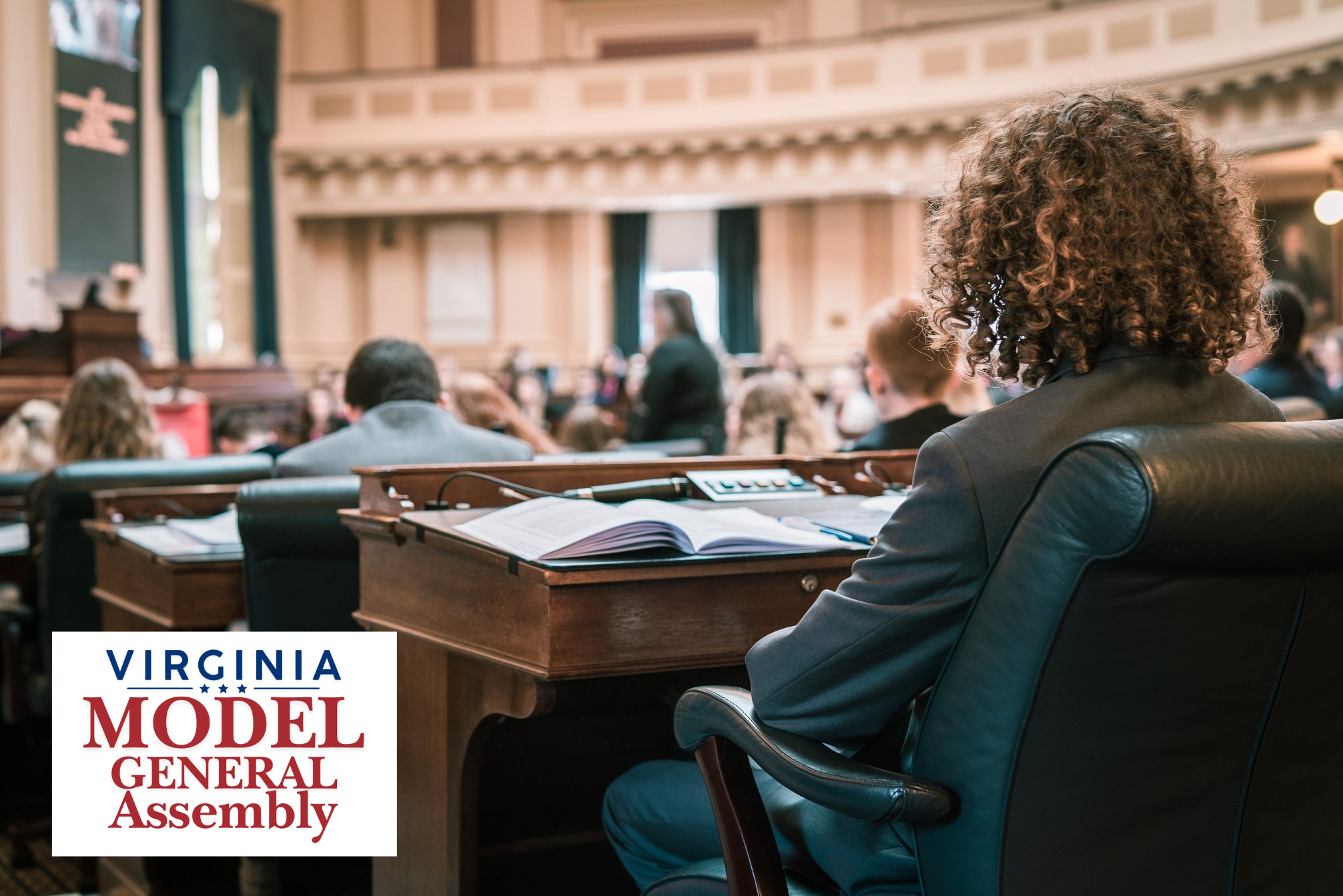 A participant viewed from behind sitting in a leather chair at a wooden desk in a grand legislative chamber, featuring the Virginia Model General Assembly logo.