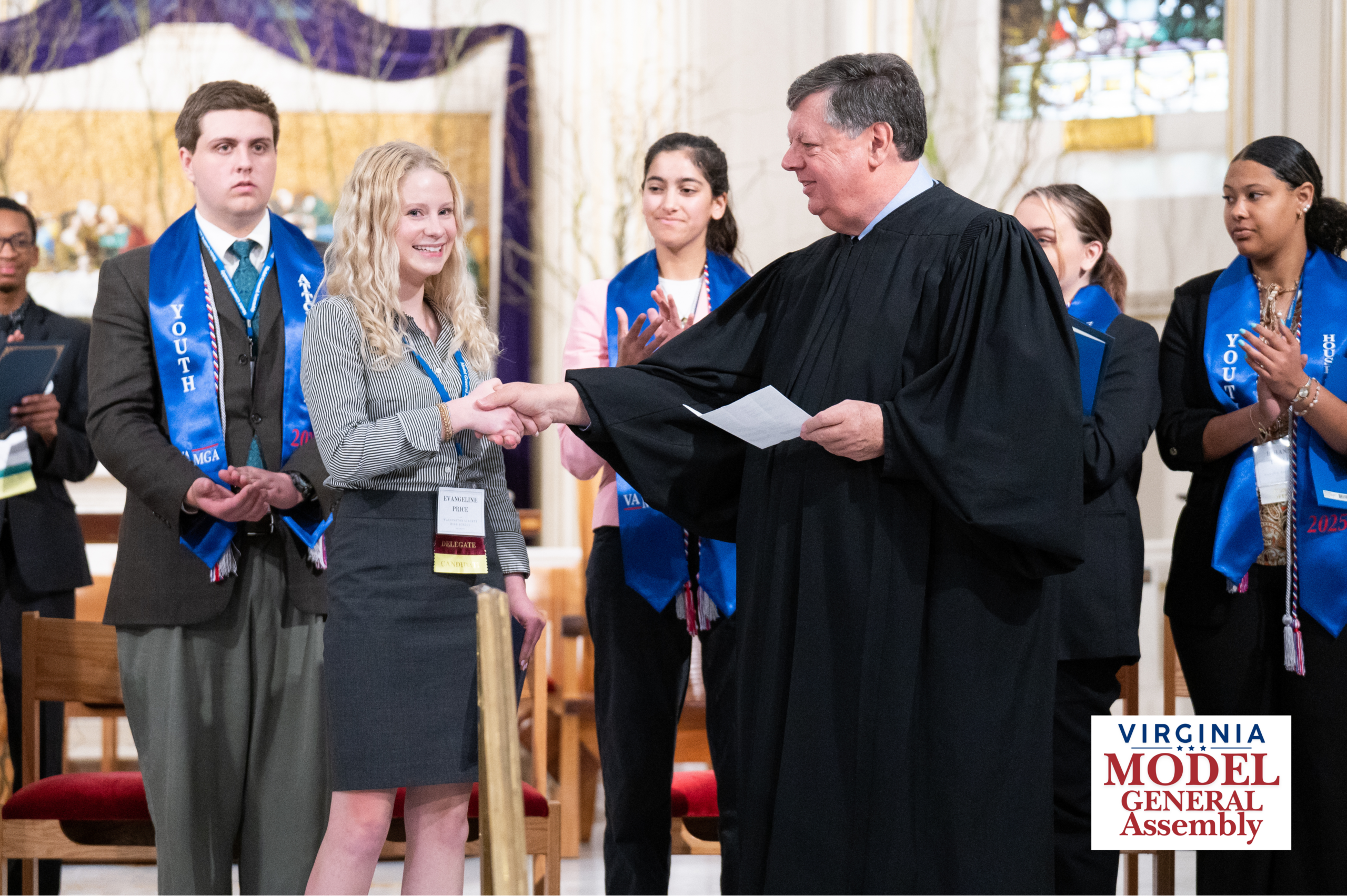 A man in a black judicial robe shaking hands with a smiling young woman on stage while other participants wearing blue sashes applaud in the background, featuring the Virginia Model General Assembly logo.