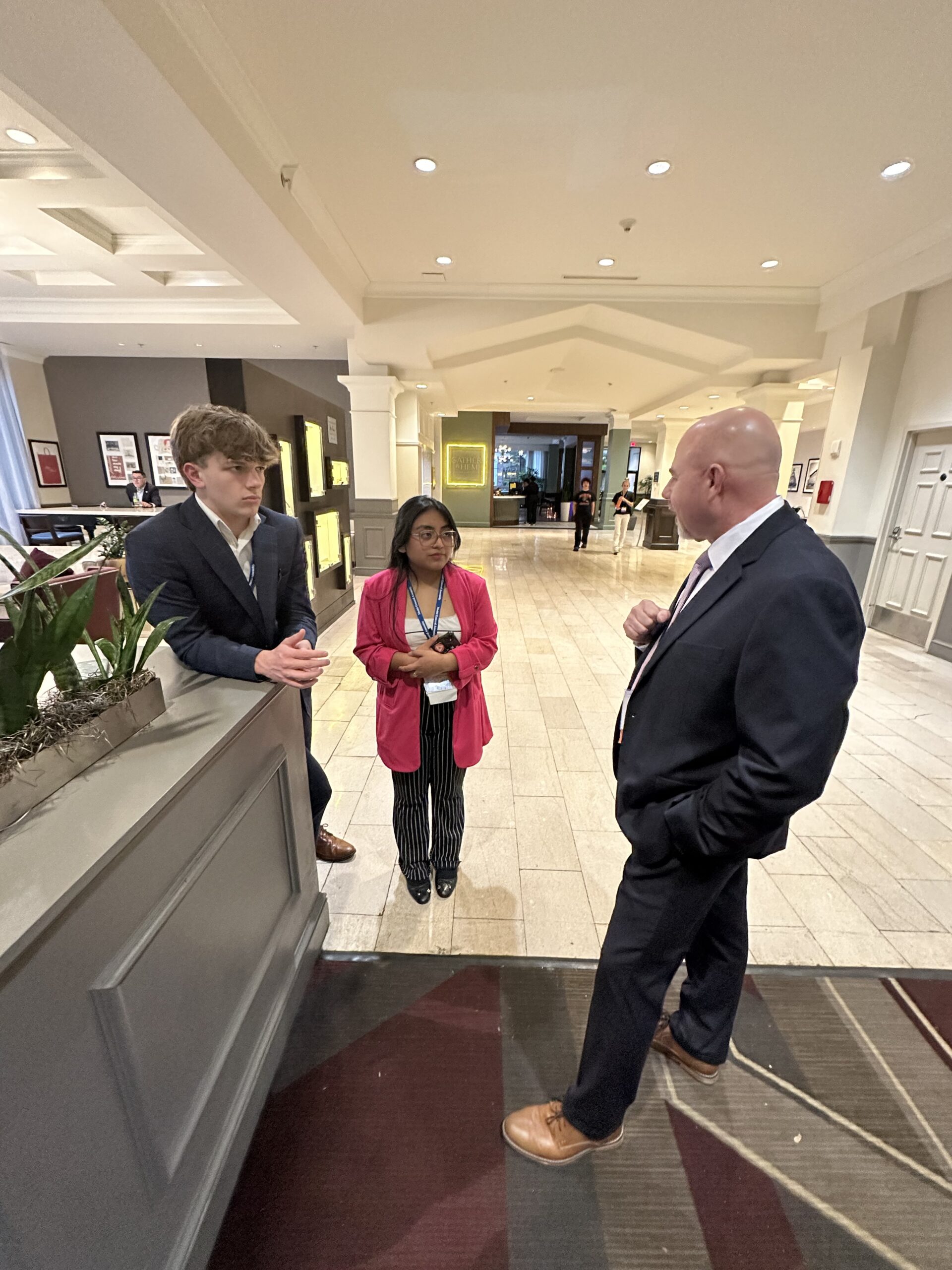 A young man and a young woman in professional attire having a conversation with an adult man in a dark suit in a hotel or convention center lobby.