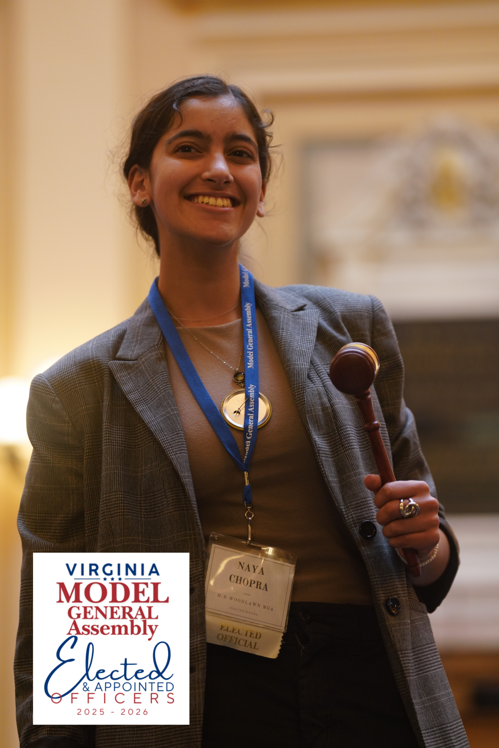 A smiling young woman holding a wooden gavel, wearing a nametag that reads 'Naya Chopra, Elected Official', featuring the Virginia Model General Assembly Elected and Appointed Officers 2025-2026 logo.