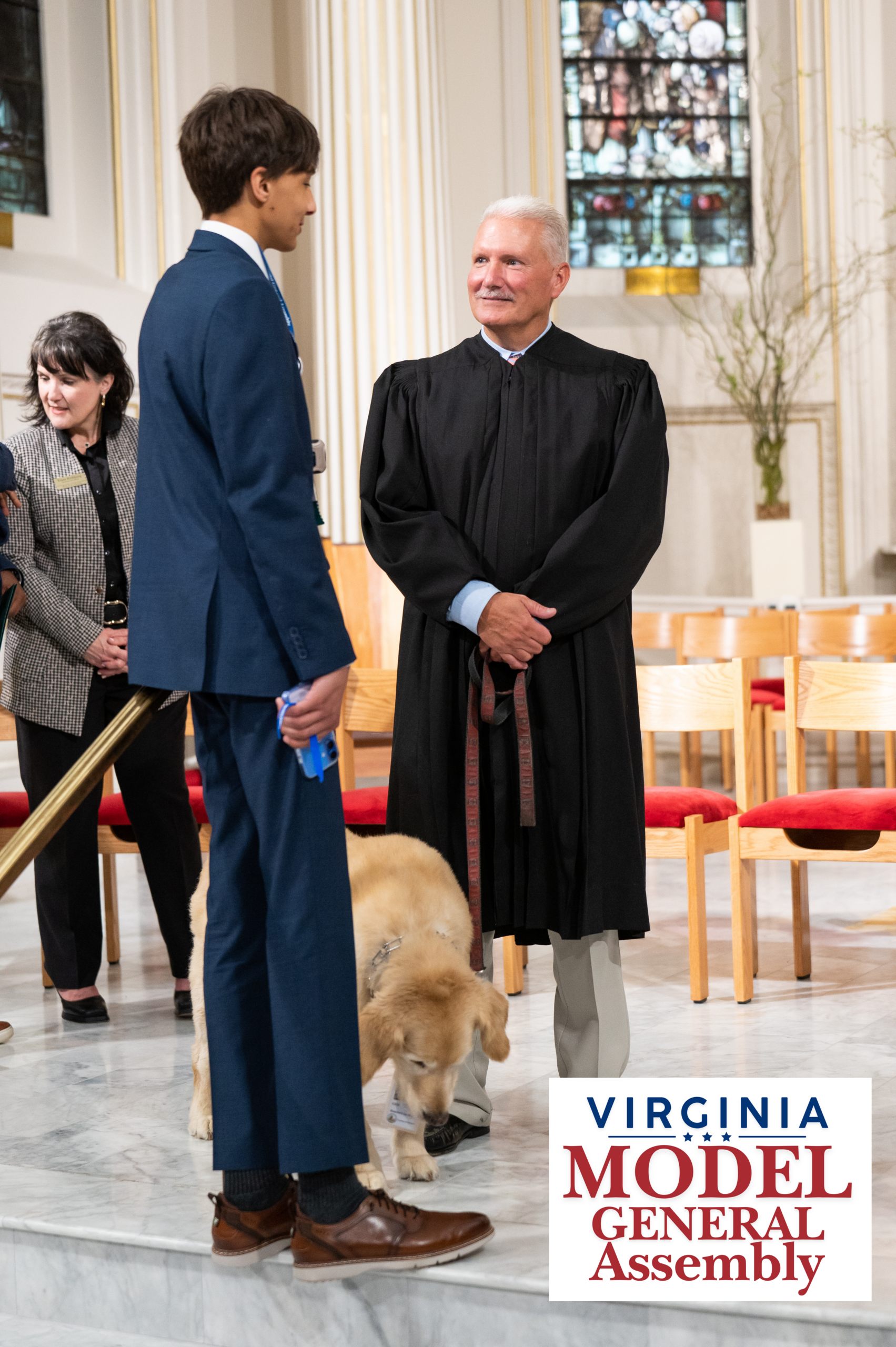 A young man in a blue suit and a man in a black judicial robe standing together near a golden retriever wearing an event lanyard, featuring the Virginia Model General Assembly logo.