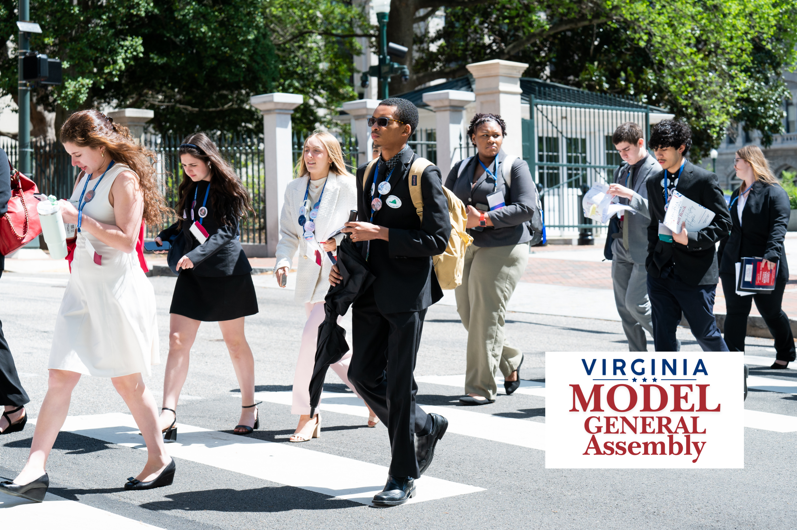 A group of young adults in professional attire and event lanyards walking across a city crosswalk on a sunny day, featuring the Virginia Model General Assembly logo.
